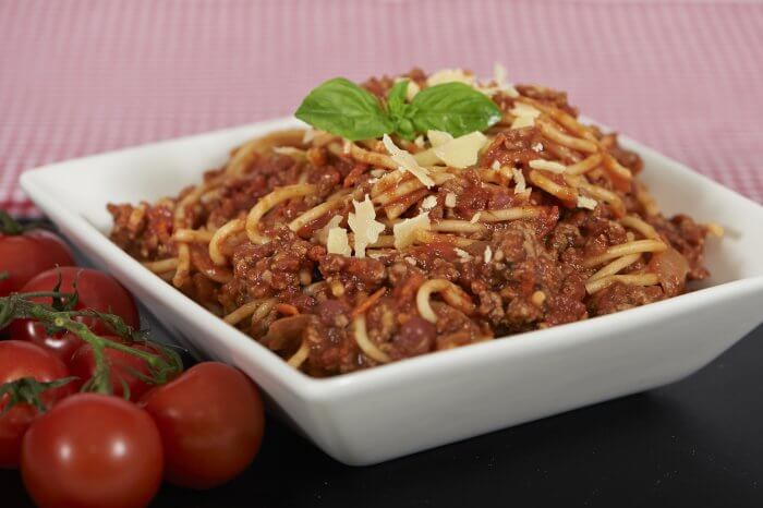 The image shows a bowl of spaghetti Bolognese topped with a rich tomato and meat sauce, garnished with Parmesan shavings and a basil leaf.